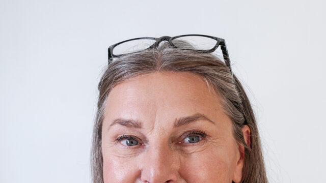 Mature woman with glasses on top of her head smiling in front of a grey background.
