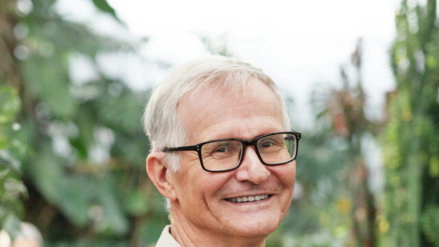 Older man in glasses smiling in front of trees.